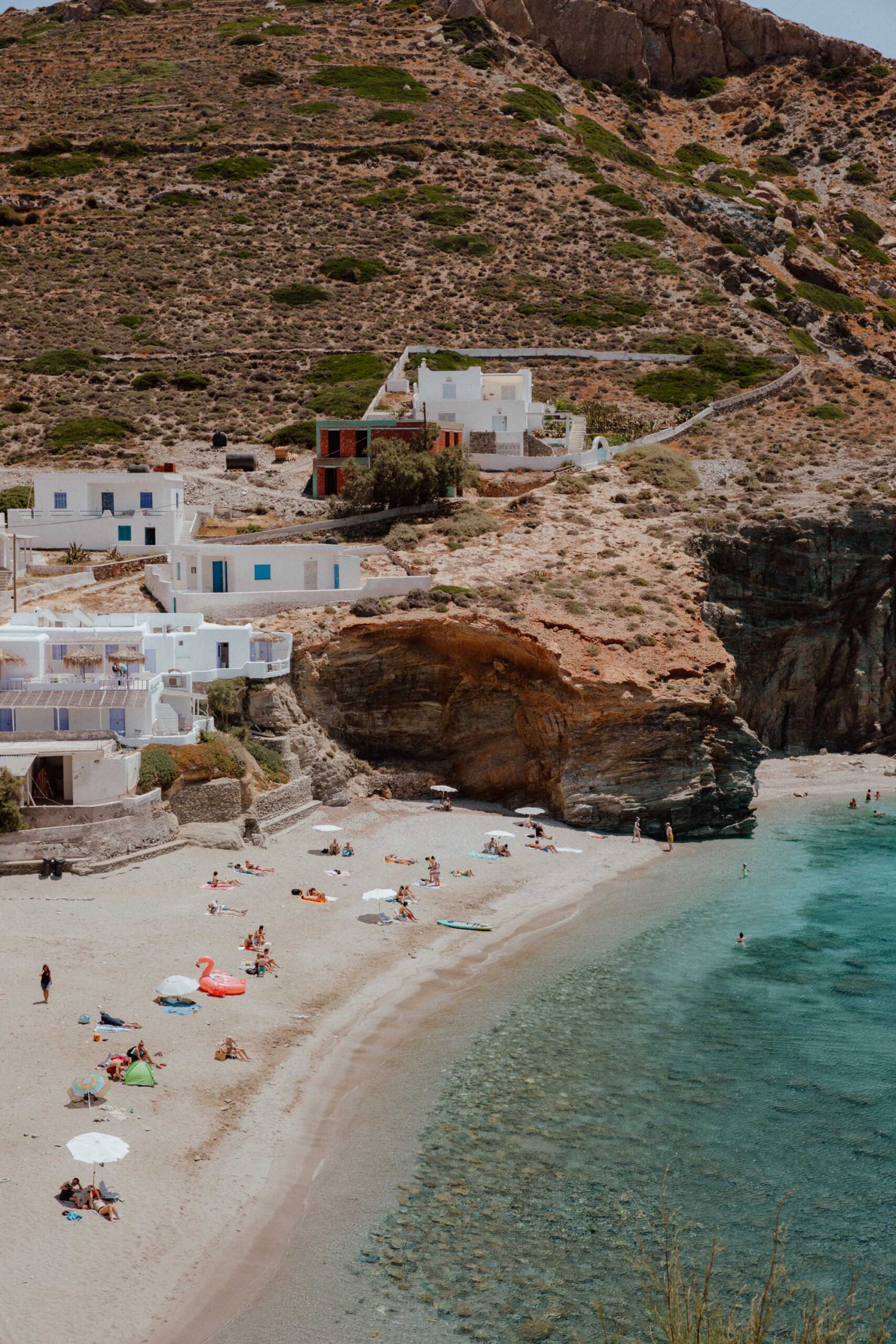 Agali Beach Folegandros with crystal clear waters, sunbathers, and whitewashed buildings nestled against dramatic cliff backdrop